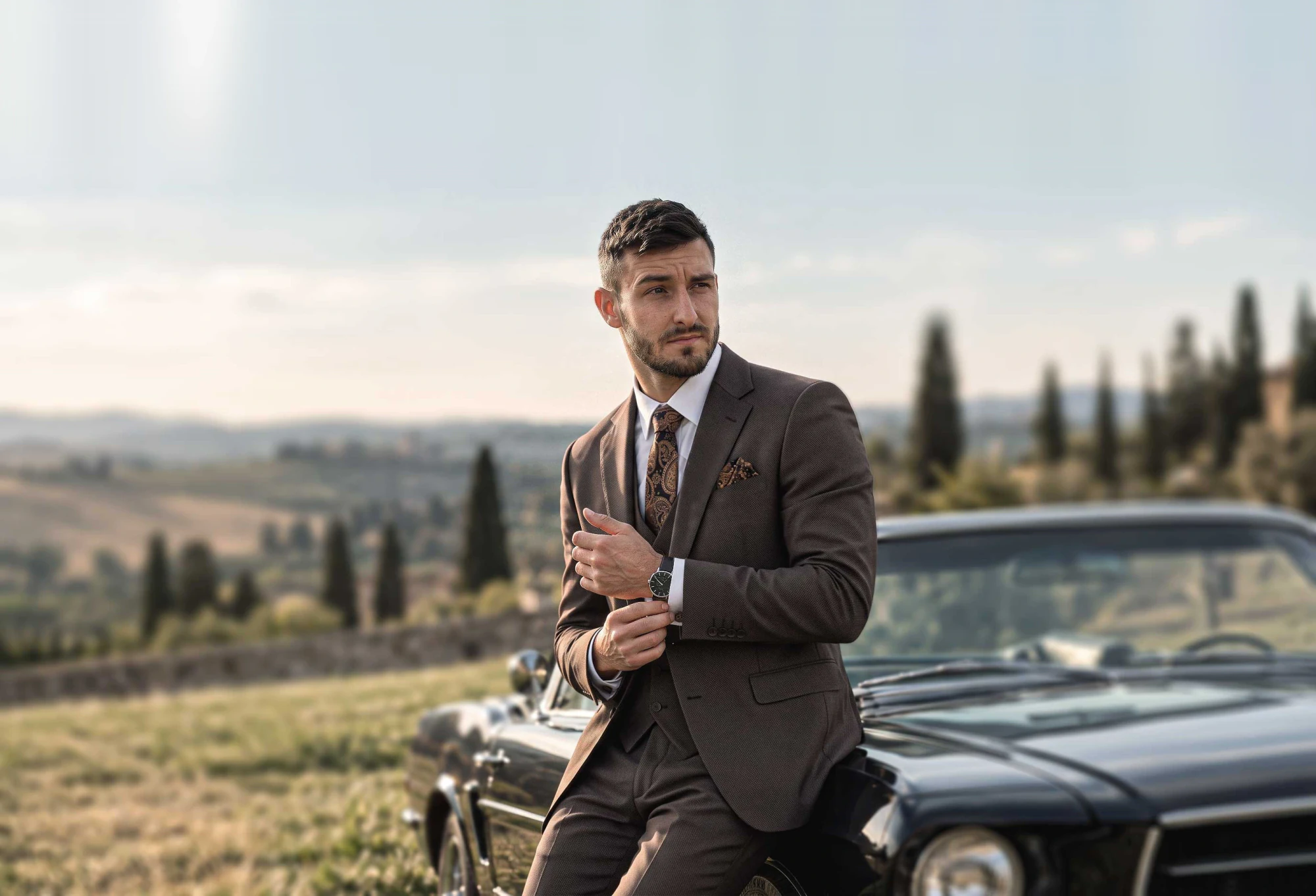 Man in een bruin maatpak bij een klassieke auto in een zuidelijk landschap